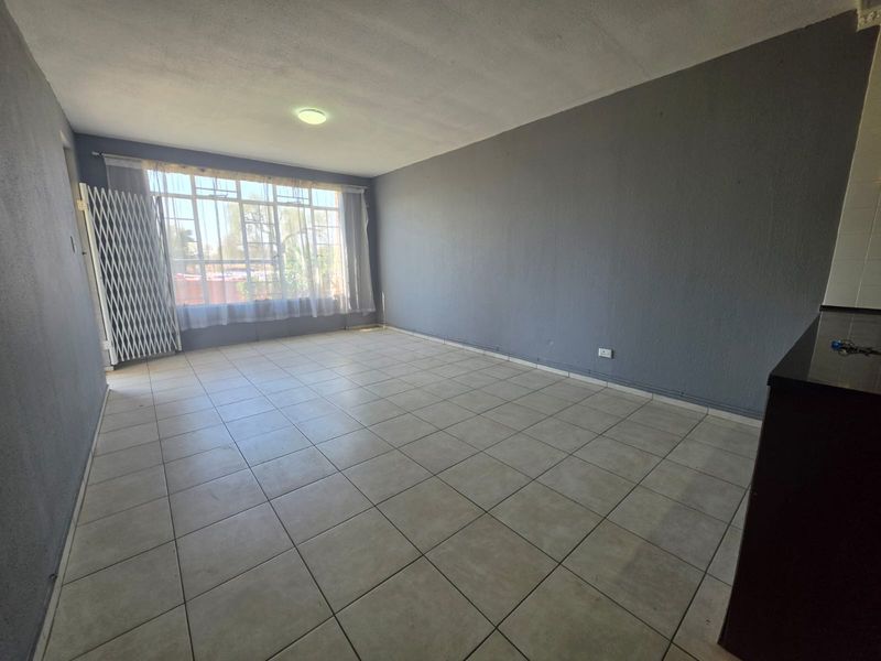 An empty open-plan living area featuring neutral tiled flooring, grey painted walls, and a large window with a security gate.