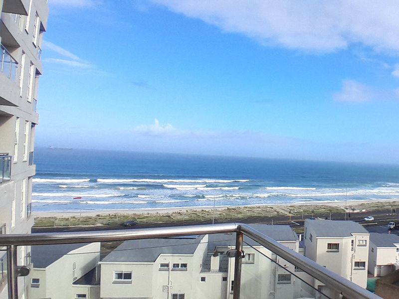 Elevated coastal view overlooking the beach and ocean, captured from an apartment balcony with a modern glass and stainless steel balustrade.