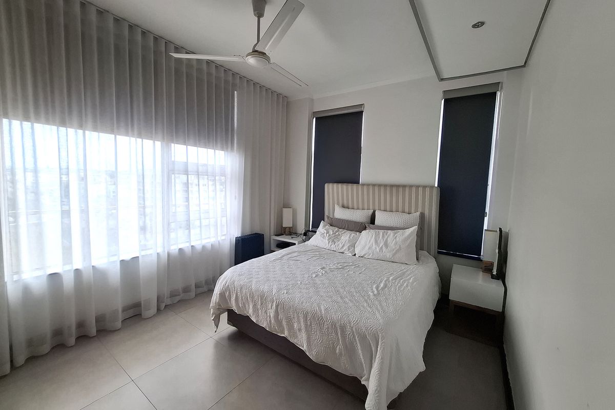A well-maintained bedroom featuring large windows with roller blinds and sheer curtains, tiled flooring, and a modern ceiling fan.