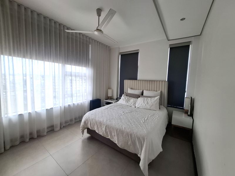 A well-maintained bedroom featuring large windows with roller blinds and sheer curtains, tiled flooring, and a modern ceiling fan.