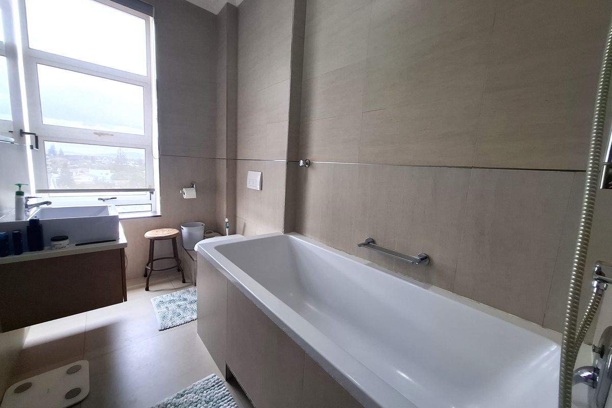 Modern bathroom featuring a built-in bathtub, wall-mounted vanity with basin, neutral tiled finishes, and a large window providing natural light.