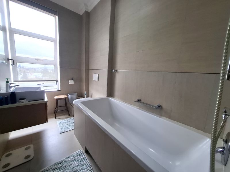 Modern bathroom featuring a built-in bathtub, wall-mounted vanity with basin, neutral tiled finishes, and a large window providing natural light.