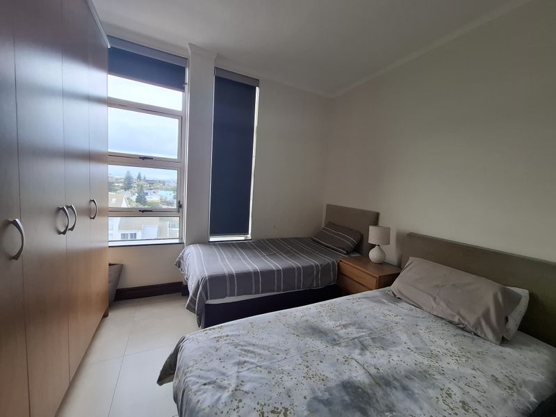 A functional bedroom featuring built-in wardrobes, tiled flooring, and large windows with roller blinds, offering natural light and a view of the surrounding area.