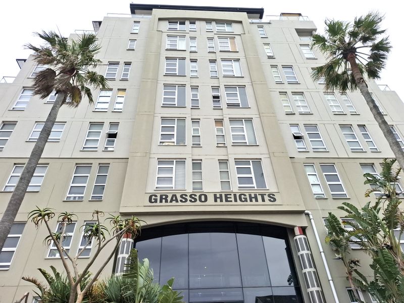 Low-angle view of the Grasso Heights apartment building facade, featuring a modern multi-story design with large glass entrance and numerous windows.