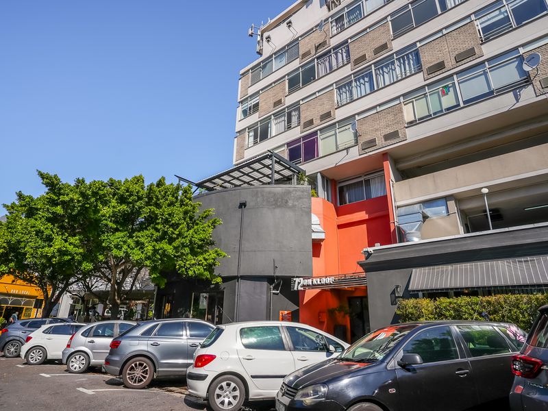 Exterior view of a multi-story apartment building facade featuring a prominent entrance, parking area, and modern architectural detailing.