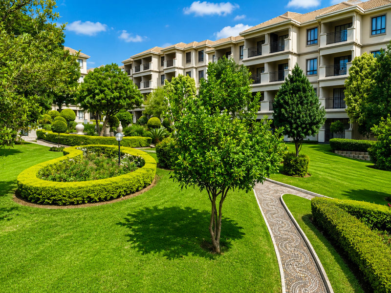 A well-maintained apartment complex featuring lush landscaped gardens, manicured hedges, and paved walkways leading to the building's rear facade with private balconies.