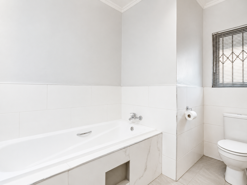 A clean, modern bathroom featuring a built-in bathtub, white ceramic wall tiles, a toilet, and a window with venetian blinds.