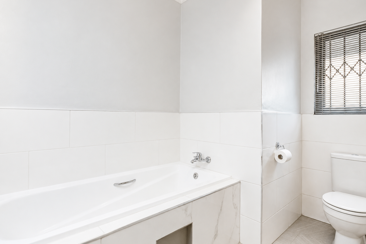 A clean, modern bathroom featuring a built-in bathtub, white ceramic wall tiles, a toilet, and a window with venetian blinds.