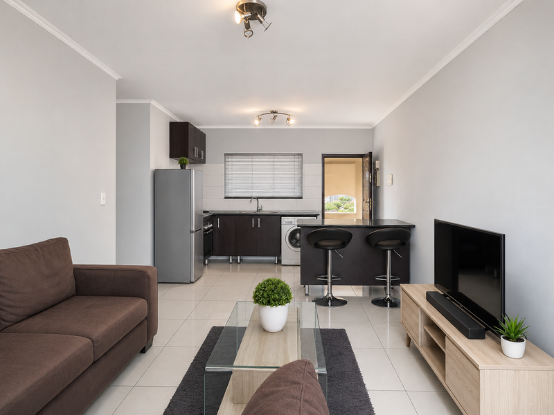 A modern open-plan living and kitchen area featuring tiled floors, dark cabinetry, a breakfast bar, and neutral wall tones.