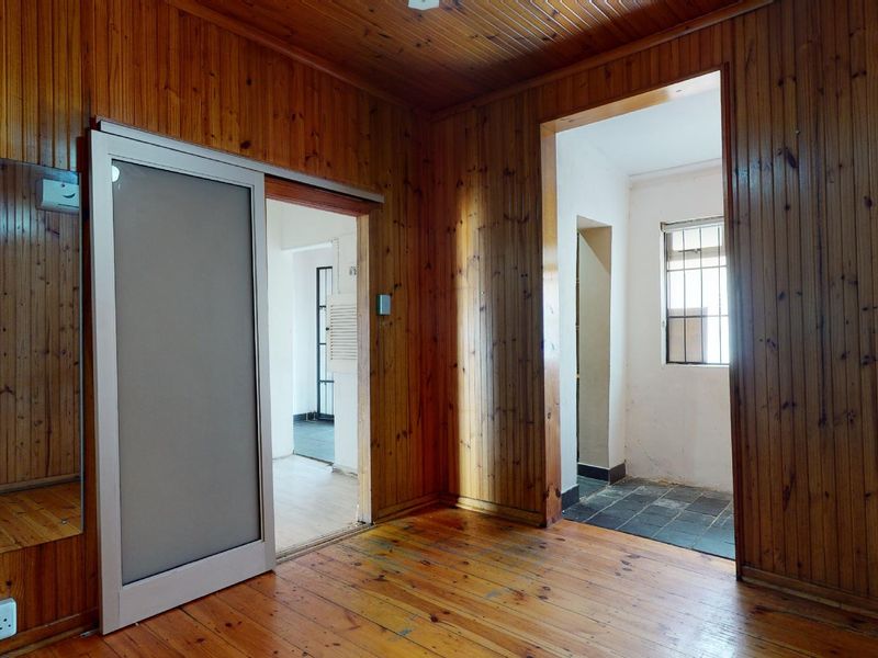 An empty living area featuring extensive timber wall paneling, wood ceilings, and timber flooring, with access to adjacent rooms and a window with security bars.