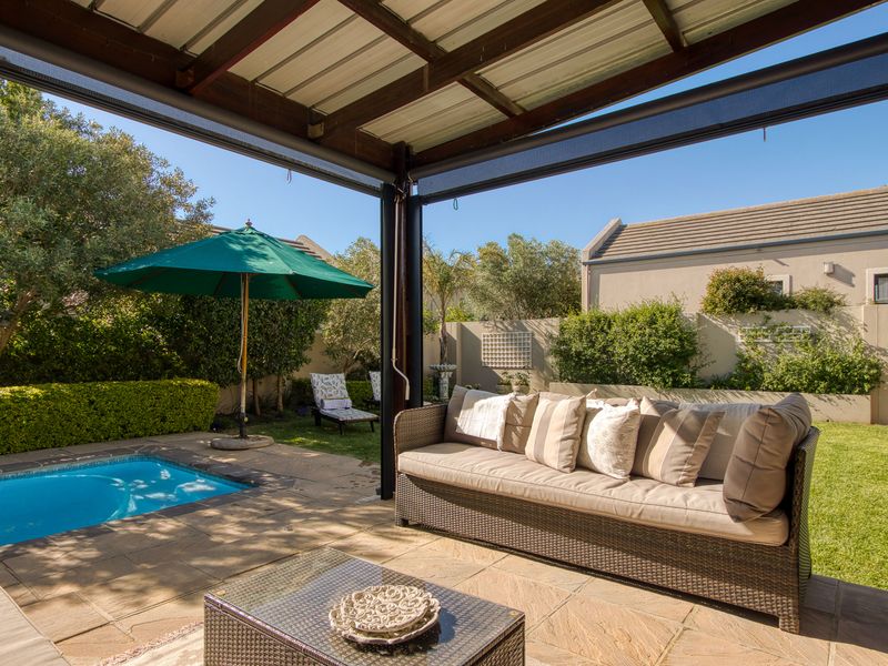 A well-maintained covered patio area featuring stone paving, overlooking a private swimming pool and a landscaped garden with boundary walls.