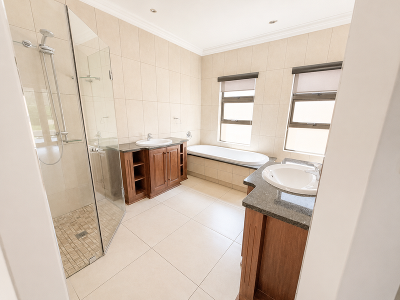 Spacious bathroom featuring a glass-enclosed shower, built-in bathtub, dual vanities with granite countertops, and neutral floor-to-ceiling tiling.