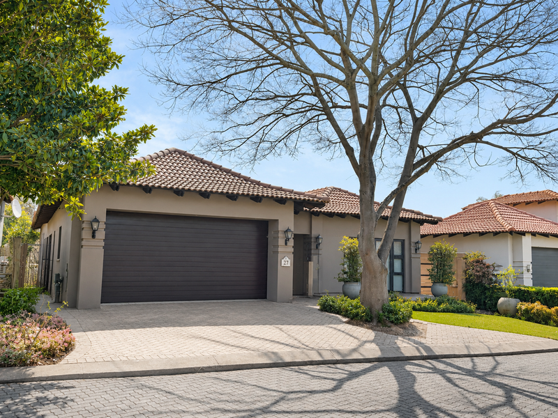 A well-maintained single-story residential property featuring a paved driveway, automated garage, and established garden with mature trees.