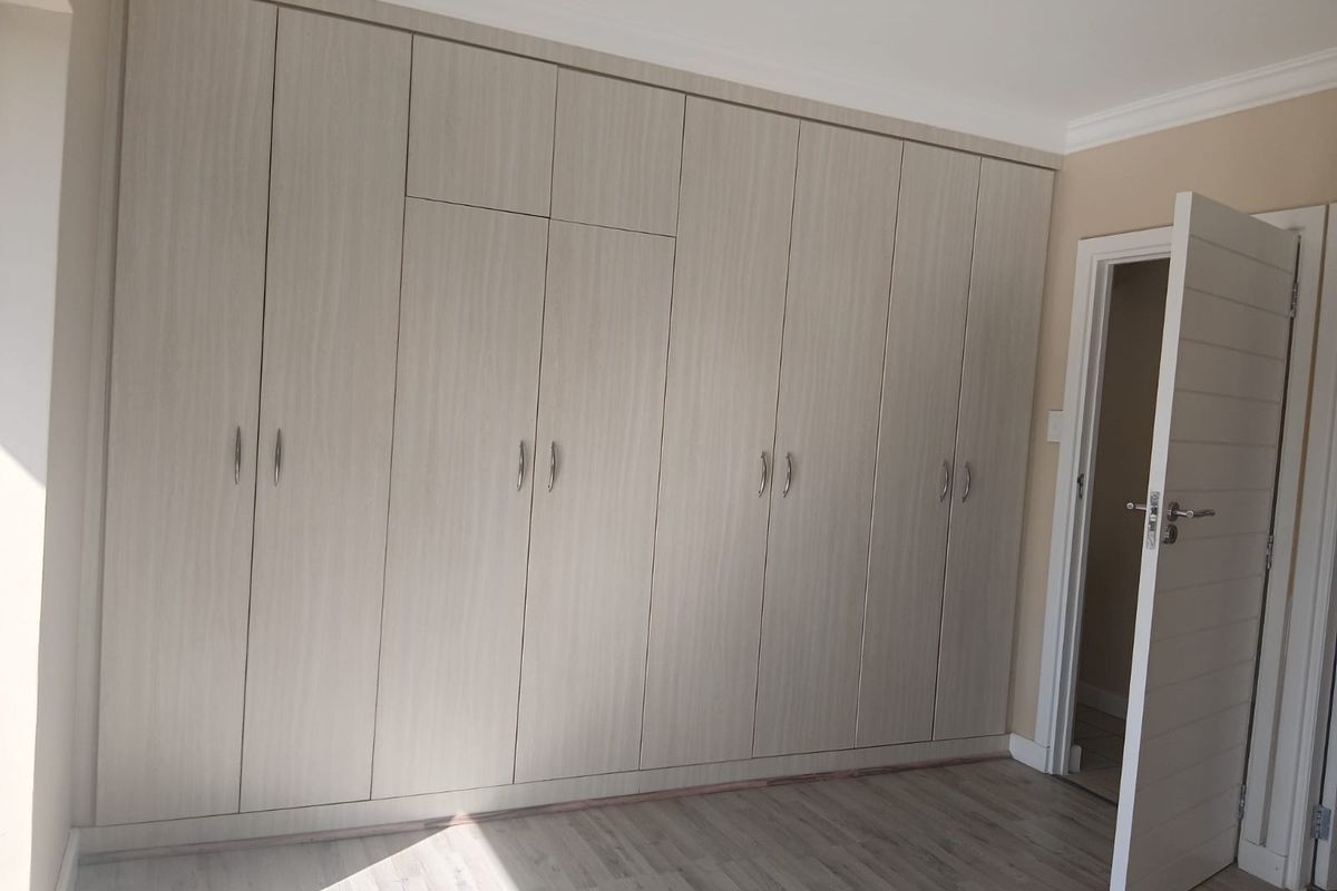 A bright bedroom featuring extensive built-in cupboards with light wood-grain finish, modern laminate flooring, and clean neutral wall finishes.