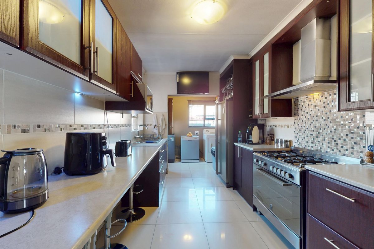 A functional galley-style kitchen featuring dark wood cabinetry, a gas hob with extractor, mosaic tile backsplash, and light-colored tiled flooring.