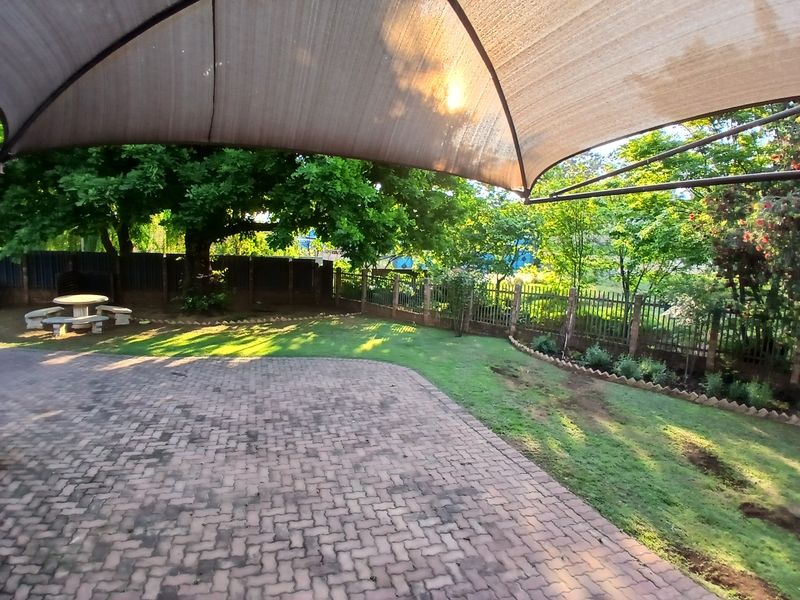A covered outdoor patio area featuring a shade sail, paved flooring, and a well-maintained garden with mature trees and perimeter fencing.
