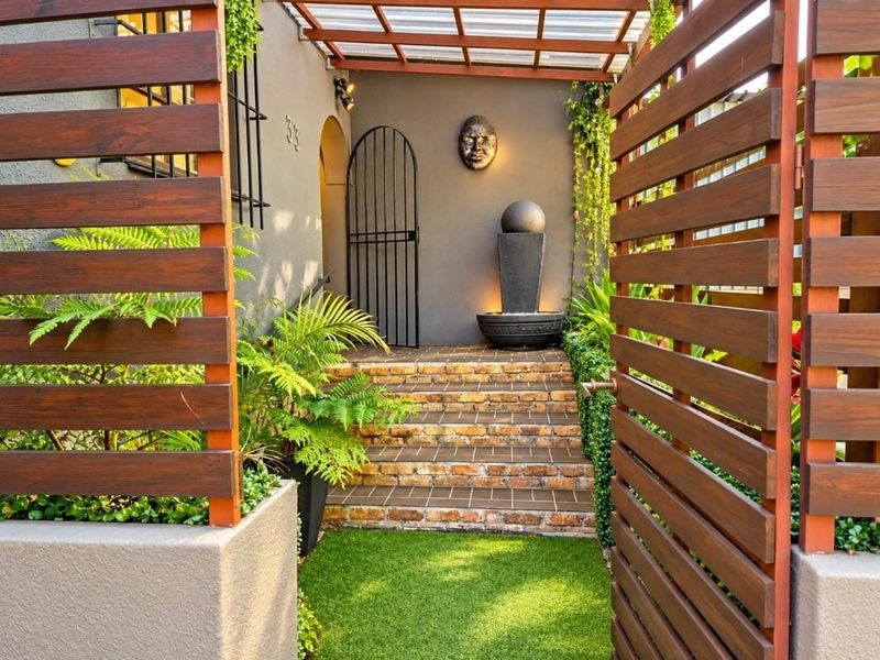 A beautifully landscaped front entrance featuring timber slat fencing, brick steps, a covered walkway, and a modern water feature.