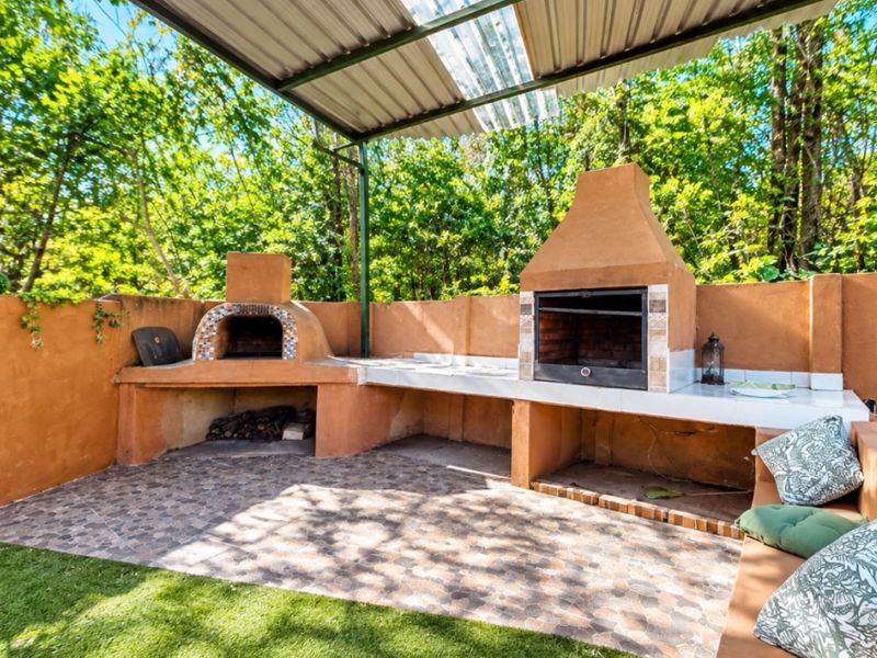 Outdoor entertainment area featuring a built-in braai, pizza oven, tiled prep surfaces, and a covered patio structure overlooking the garden.