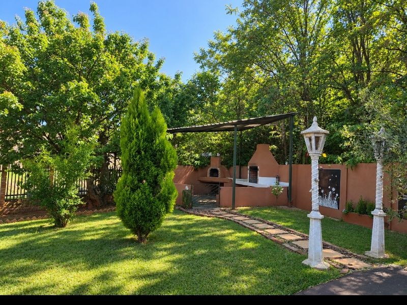 A well-maintained outdoor entertainment area featuring a built-in braai, covered patio space, and a stone pathway set within a lush, landscaped garden.