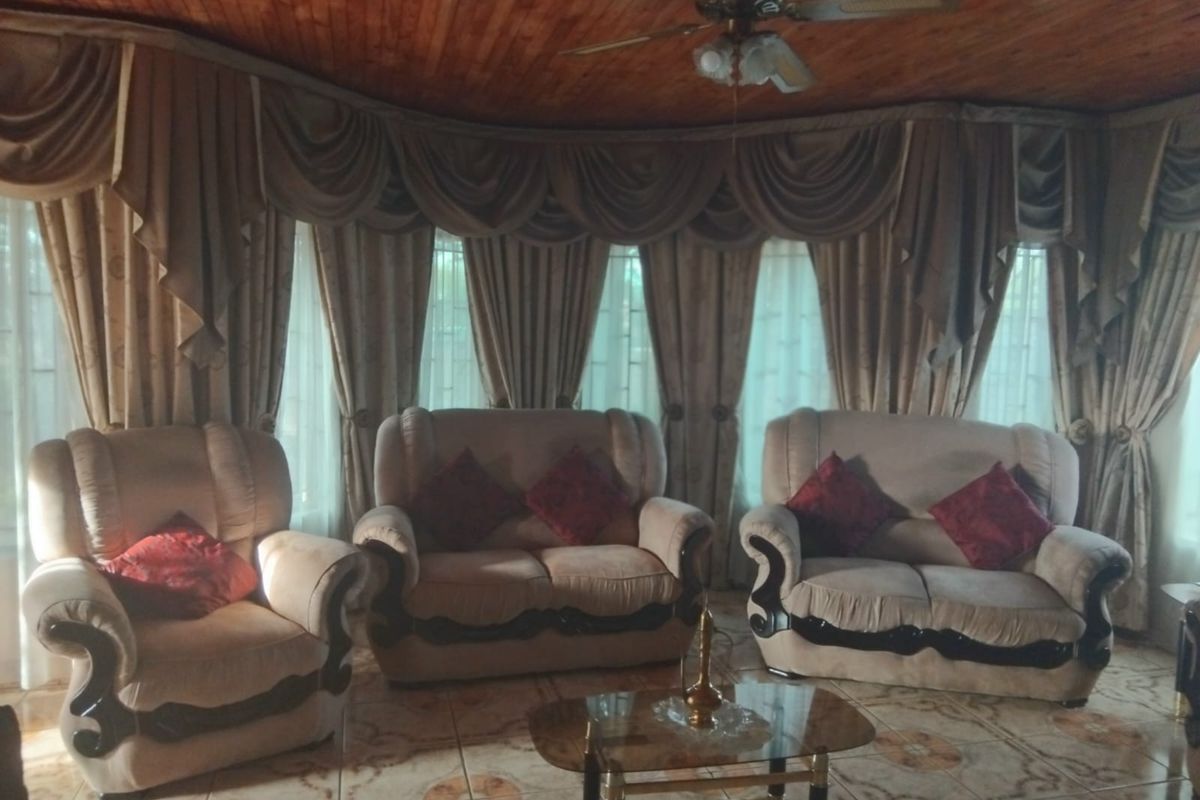 A living room featuring a timber tongue and groove ceiling, ceiling fan, and ceramic floor tiles, with large windows draped in heavy curtains.