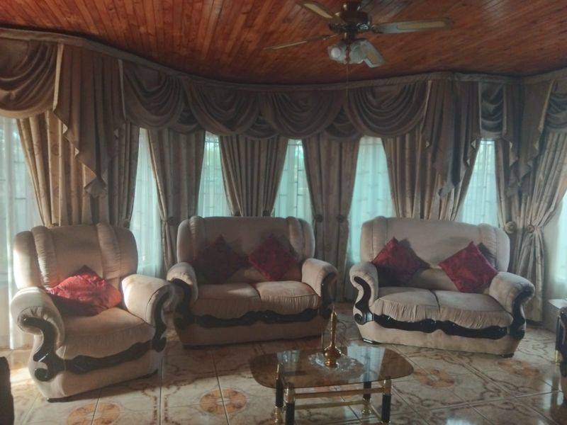 A living room featuring a timber tongue and groove ceiling, ceiling fan, and ceramic floor tiles, with large windows draped in heavy curtains.