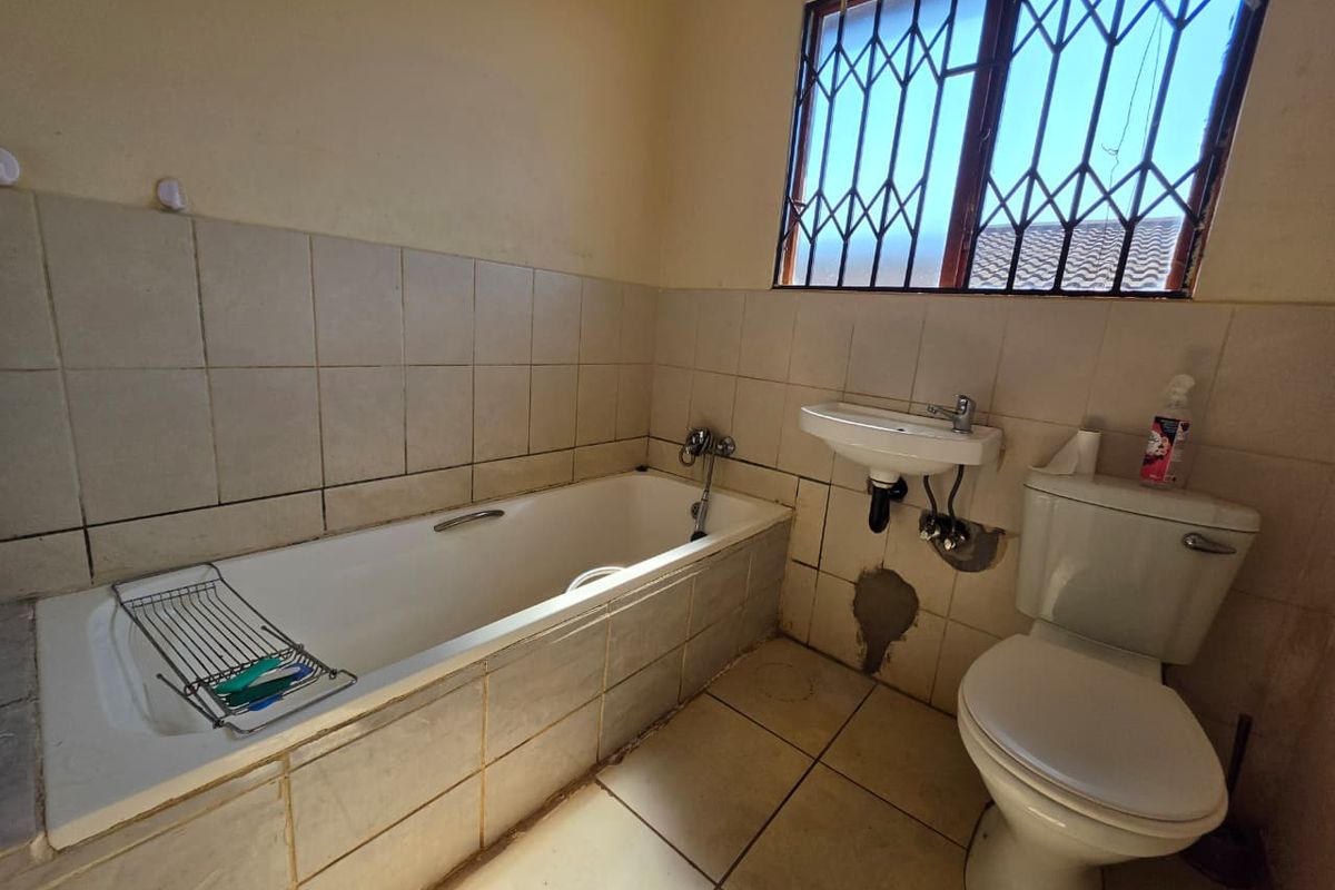 A functional bathroom featuring a built-in bathtub, wall-mounted basin, and toilet, requiring cosmetic updates and repairs to wall tiling and finishes.
