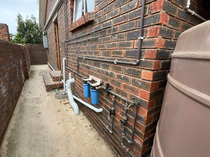 Side exterior view showing brick wall construction, external water filtration system, plumbing infrastructure, and a large water storage tank on a concrete path.