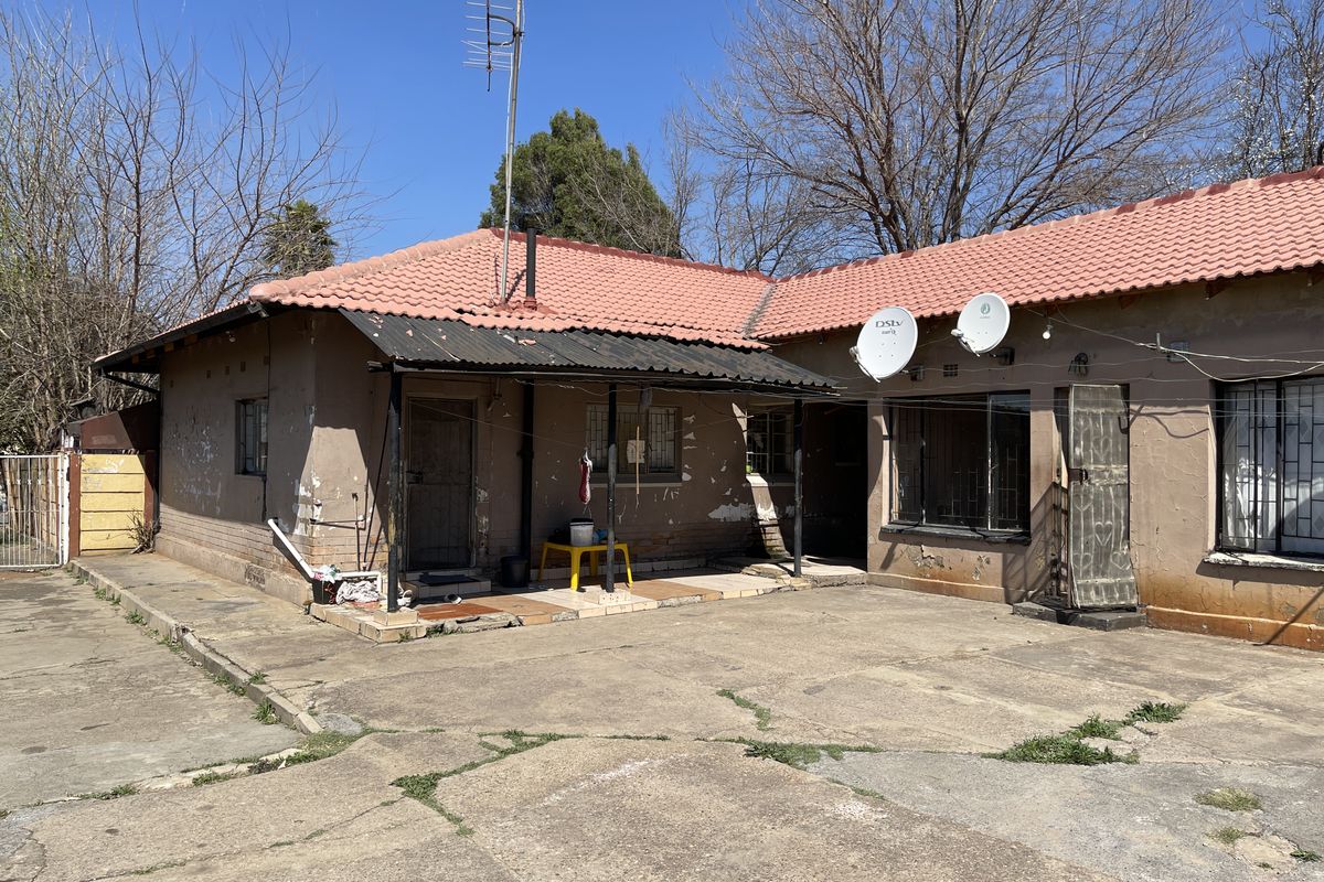 Rear exterior view of a single-story house featuring a concrete driveway, covered patio area, and tiled roof, showing signs of significant exterior wear.
