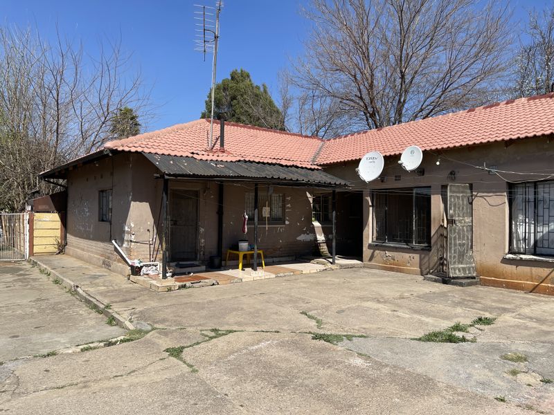 Rear exterior view of a single-story house featuring a concrete driveway, covered patio area, and tiled roof, showing signs of significant exterior wear.