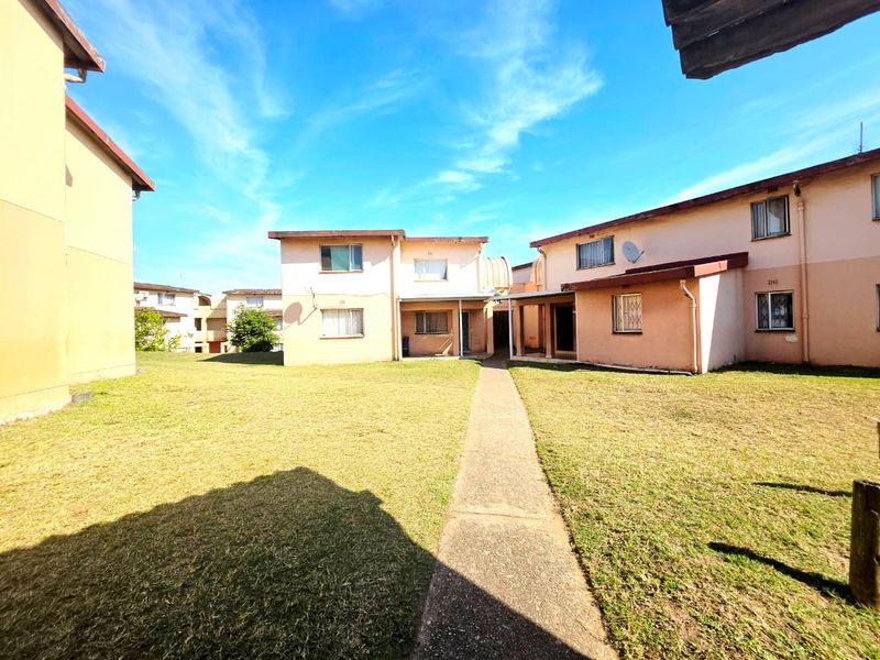 Exterior view of an apartment complex featuring a central concrete walkway, communal lawn area, and multi-story residential units with security window bars.
