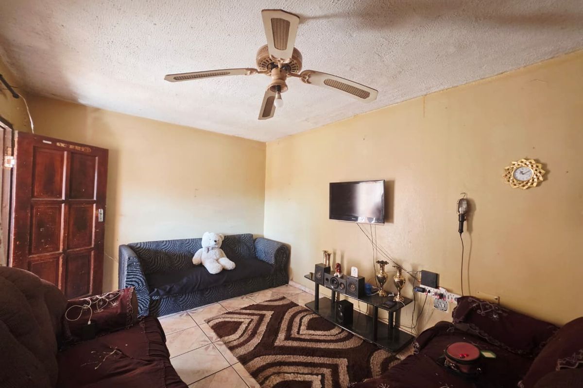 A living room featuring tiled flooring, a ceiling fan, and a solid wood door, currently requiring cosmetic updates to the walls and ceiling.