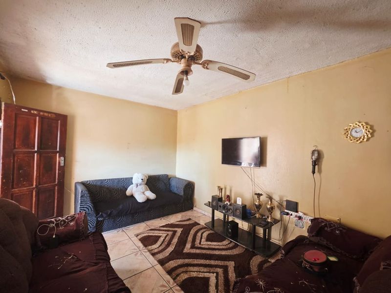 A living room featuring tiled flooring, a ceiling fan, and a solid wood door, currently requiring cosmetic updates to the walls and ceiling.