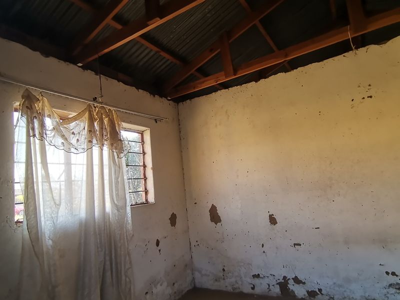An empty bedroom featuring exposed timber roof trusses, corrugated metal roofing, and walls requiring significant plaster and paint restoration.