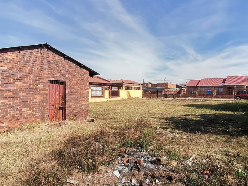 A view of the rear yard featuring a brick outbuilding, perimeter fencing, and an open grass area requiring landscaping.