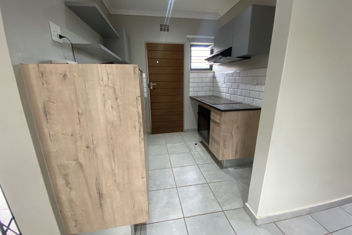 A modern, compact kitchen featuring wood-look cabinetry, a built-in oven and hob, subway tile backsplash, and neutral tiled flooring.