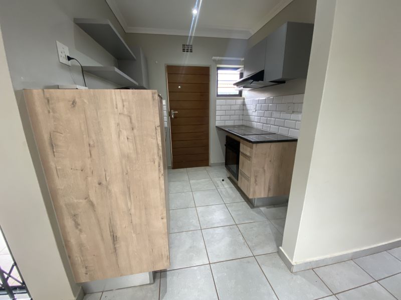 A modern, compact kitchen featuring wood-look cabinetry, a built-in oven and hob, subway tile backsplash, and neutral tiled flooring.
