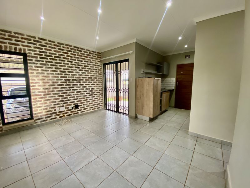 Open plan living area featuring a striking exposed brick wall, neutral floor tiles, recessed lighting, and a compact kitchen area with modern cabinetry.