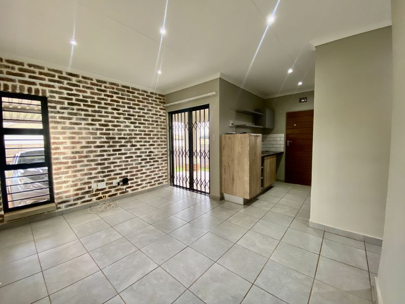Open-plan living area featuring a striking exposed brick wall, modern tiled flooring, recessed lighting, and a compact built-in kitchenette.