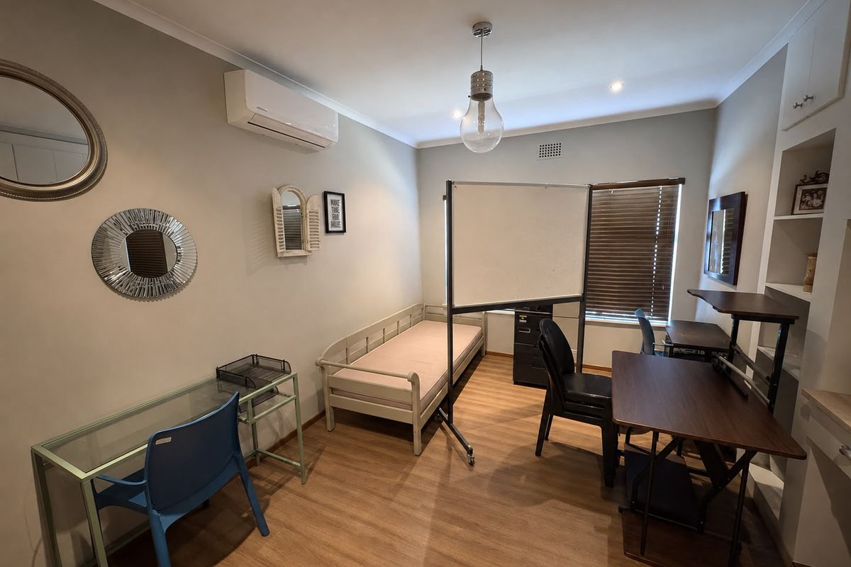 A functional bedroom featuring laminate flooring, built-in shelving, and an air conditioning unit, currently utilized as a multi-purpose study and living space.