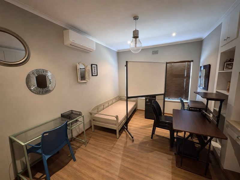 A functional bedroom featuring laminate flooring, built-in shelving, and an air conditioning unit, currently utilized as a multi-purpose study and living space.