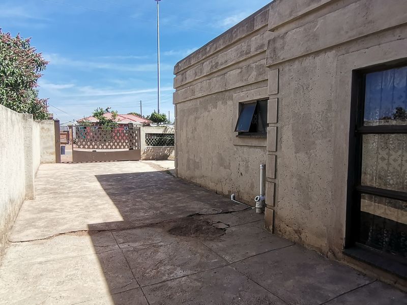 Rear exterior view showing a paved driveway area, boundary wall with metal gate, and weathered exterior wall finish.