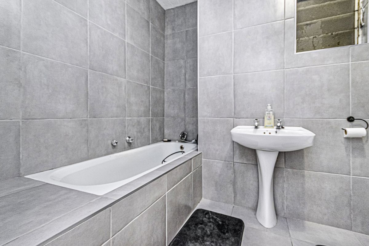 A compact bathroom featuring grey wall and floor tiles, a built-in bathtub, and a white pedestal basin.