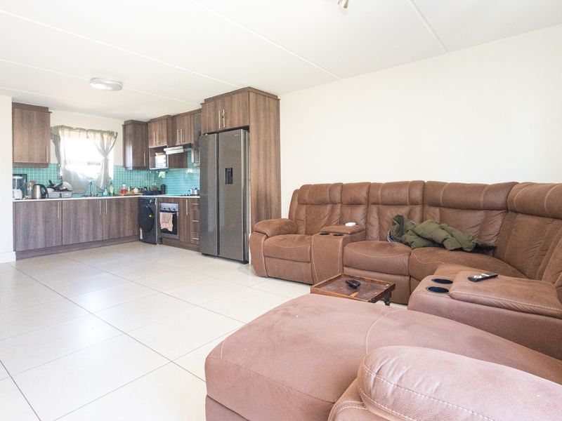 Open-plan living area featuring a modern kitchen with wood-finish cabinetry, tiled backsplash, and integrated appliances, flowing into a spacious lounge with tiled flooring.
