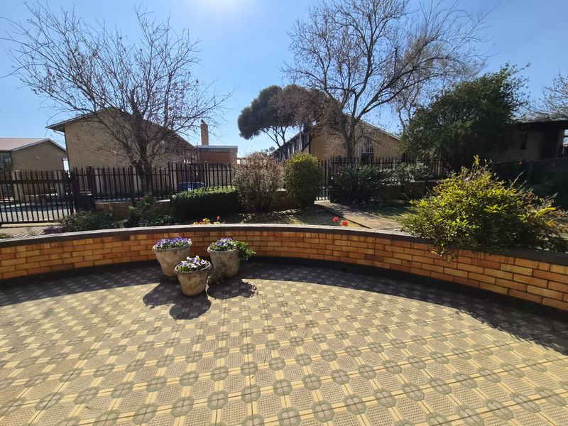 A paved outdoor patio area featuring a curved brick retaining wall and metal perimeter fencing, set within a landscaped garden environment.