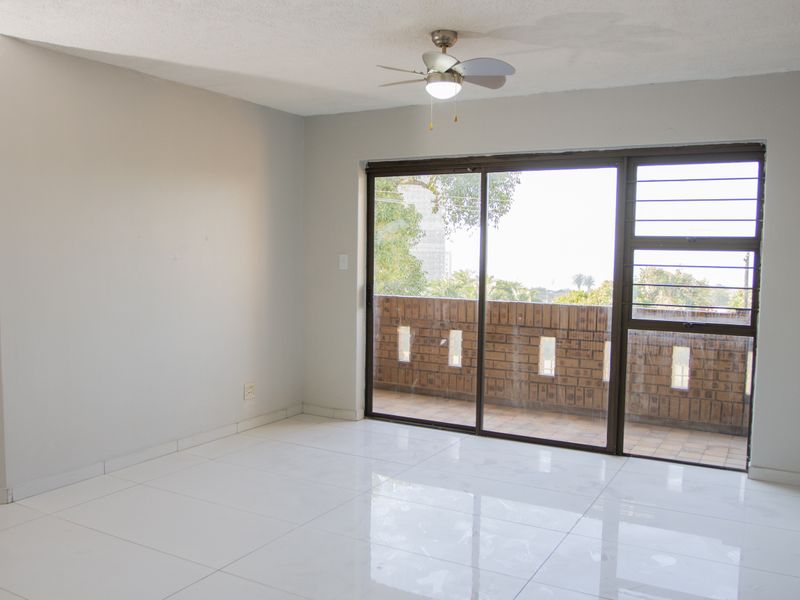 An empty living room featuring white tiled flooring, a ceiling fan, and large sliding glass doors leading to a private balcony.