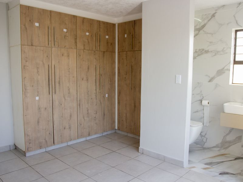 Modern bedroom featuring extensive built-in wooden cupboards, neutral tiled flooring, and a sleek en-suite bathroom with marble-look wall tiles and contemporary fixtures.
