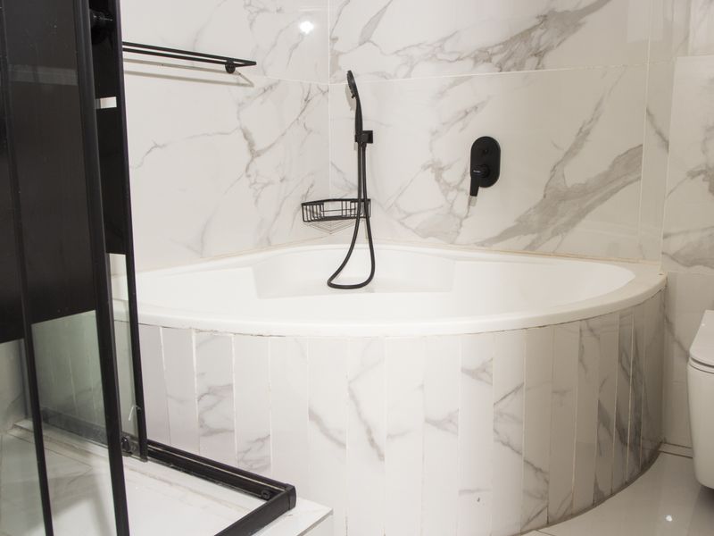 Modern bathroom featuring a corner bathtub with marble-look tiling, black fixtures, and a wall-mounted toilet.