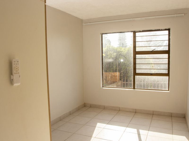 A bright, empty bedroom featuring neutral wall finishes, tiled flooring, and a large window with an aluminium frame.