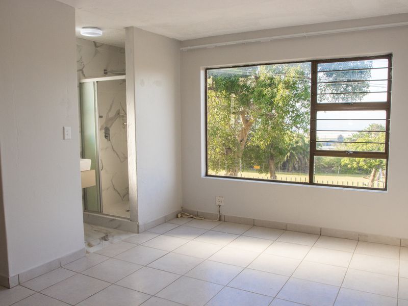 An empty bedroom featuring tiled flooring, a large window with security bars, and an integrated en-suite shower room with marble-look wall tiles.