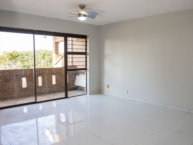 A bright, empty living room featuring white tiled flooring, a ceiling fan, and large sliding glass doors leading to an outdoor balcony area.
